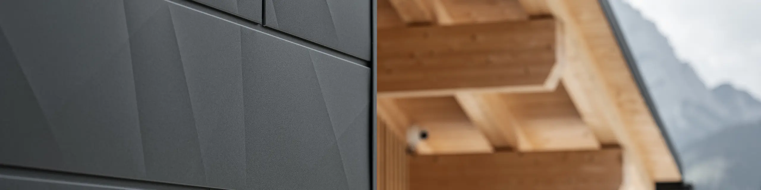 Close-up of PREFA Siding.X façade panels in P.10 black on Casa Hoffe in Sappada, Italy. The aluminium façade system is skillfully installed in a modern, abstract design, creating an appealing contrast with the visible wooden elements of the roof structure. The innovative façade solution blends seamlessly into the alpine setting and represents a combination of functionality, durability and aesthetic building culture.