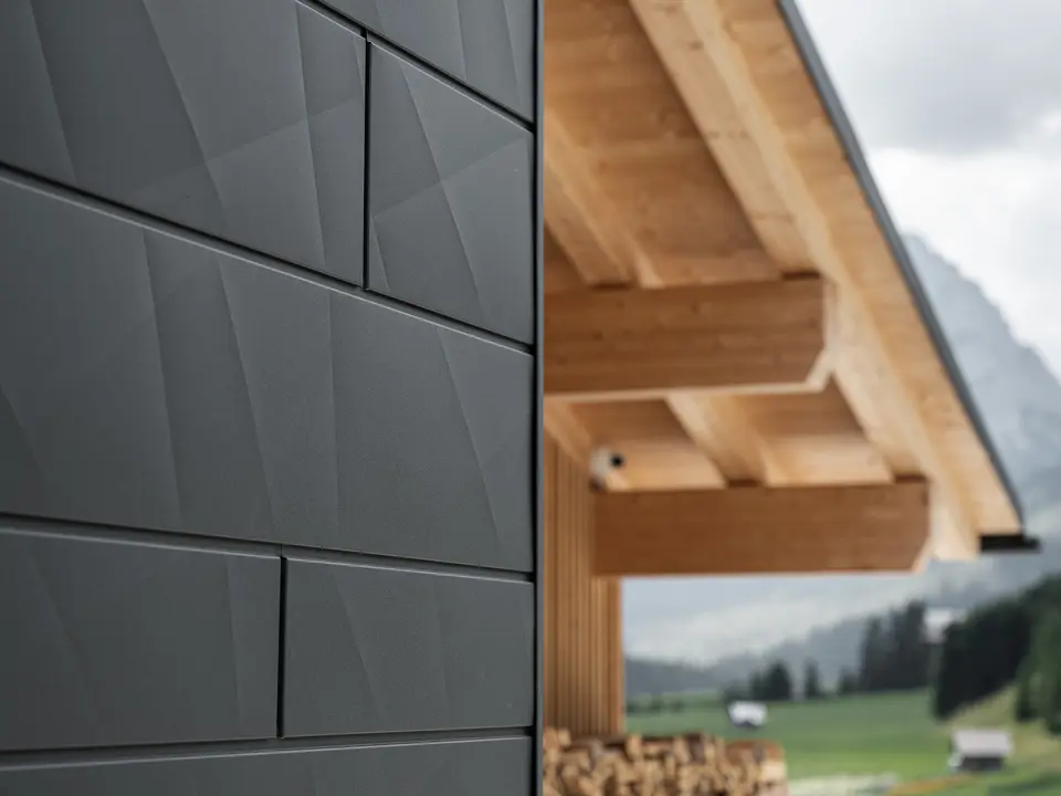 Close-up of PREFA Siding.X façade panels in P.10 black on Casa Hoffe in Sappada, Italy. The aluminium façade system is skillfully installed in a modern, abstract design, creating an appealing contrast with the visible wooden elements of the roof structure. The innovative façade solution blends seamlessly into the alpine setting and represents a combination of functionality, durability and aesthetic building culture.