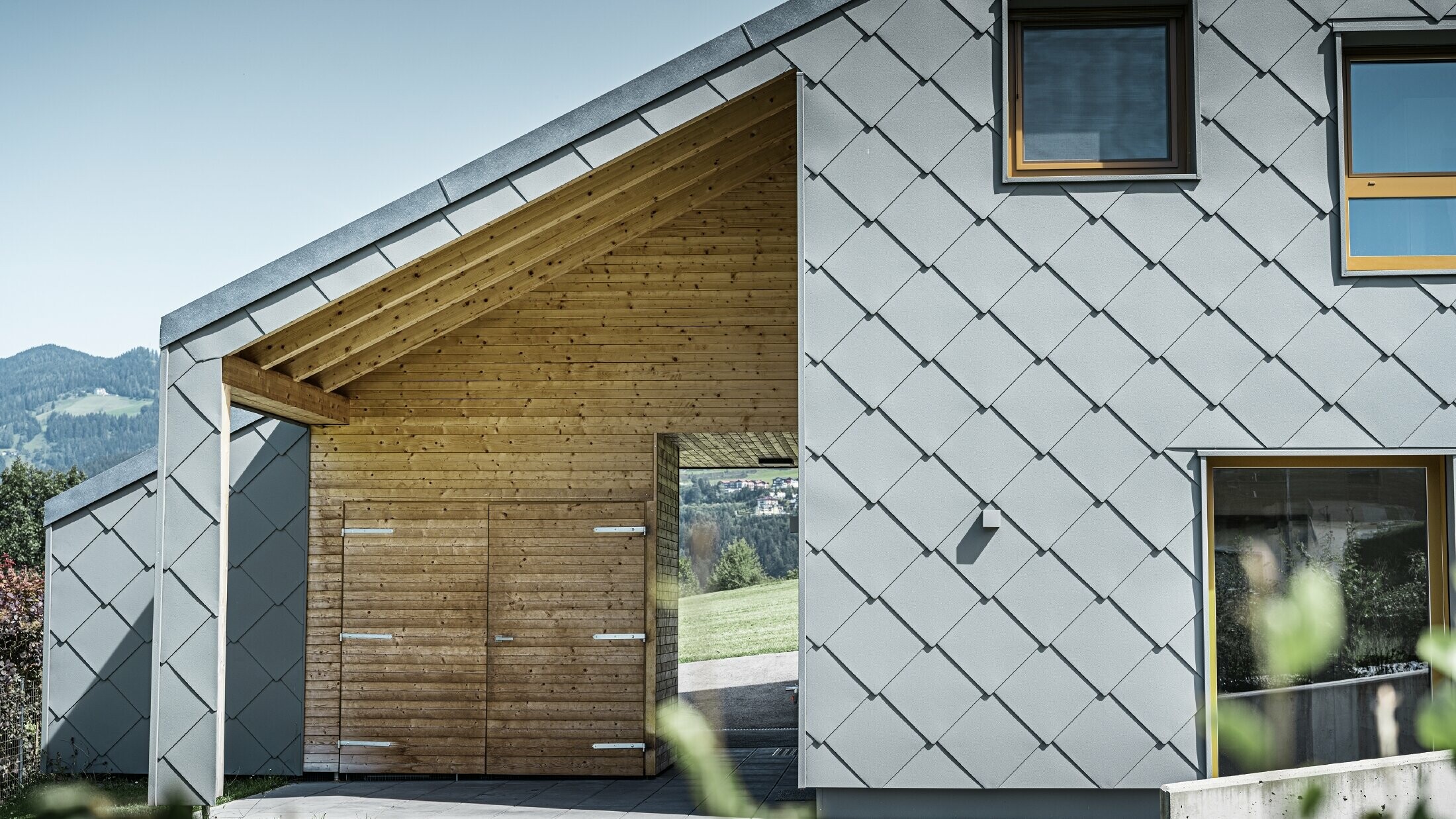 The covered entrance area is made with horizontal wood panelling, the rest of the façade is clad with the large PREFA aluminium rhomboid façade tiles in light grey