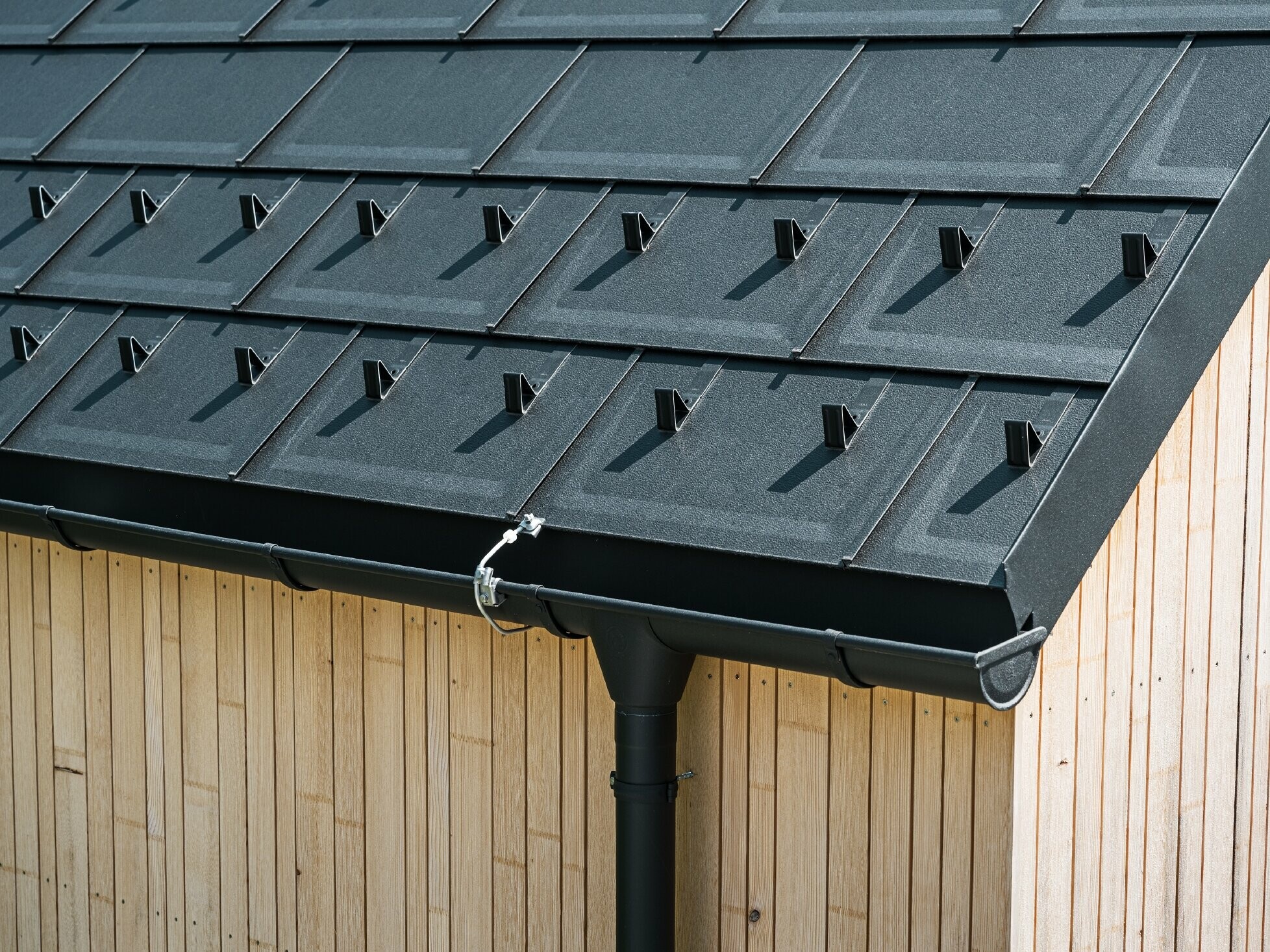 Close-up of PREFA roof tile R.16 with snow guards, gutter and downpipe in black P.10 incl. lightning protection and a vertical wooden façade