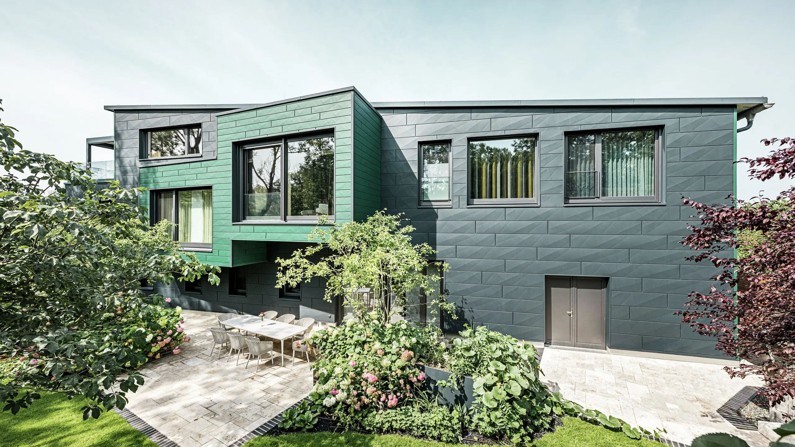 The detached house in Türkheim impresses with its unusual façade, which is clad with PREFA Siding X panels in anthracite and moss green. The clean lines and striking colour choice of the aluminium panels give the building a modern and elegant look. In the foreground is the spacious garden area with a stylish terrace and a dining table with modern chairs. Colourful flower beds and healthy plants provide a lively, green contrast to the robust façade. The durable and low-maintenance aluminium façade protects the building and ensures timeless beauty.