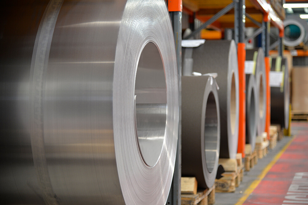 Aluminum coils stored on wooden pallets, with a plain aluminum coil in the foreground and further aluminum coils and sheets behind it.