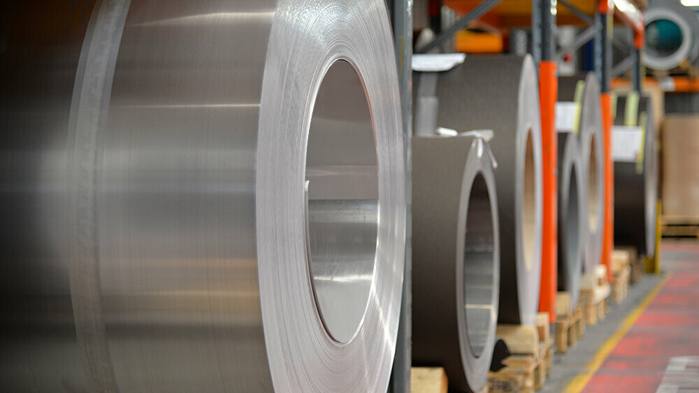 Aluminum coils stored on wooden pallets, with a plain aluminum coil in the foreground and further aluminum coils and sheets behind it.