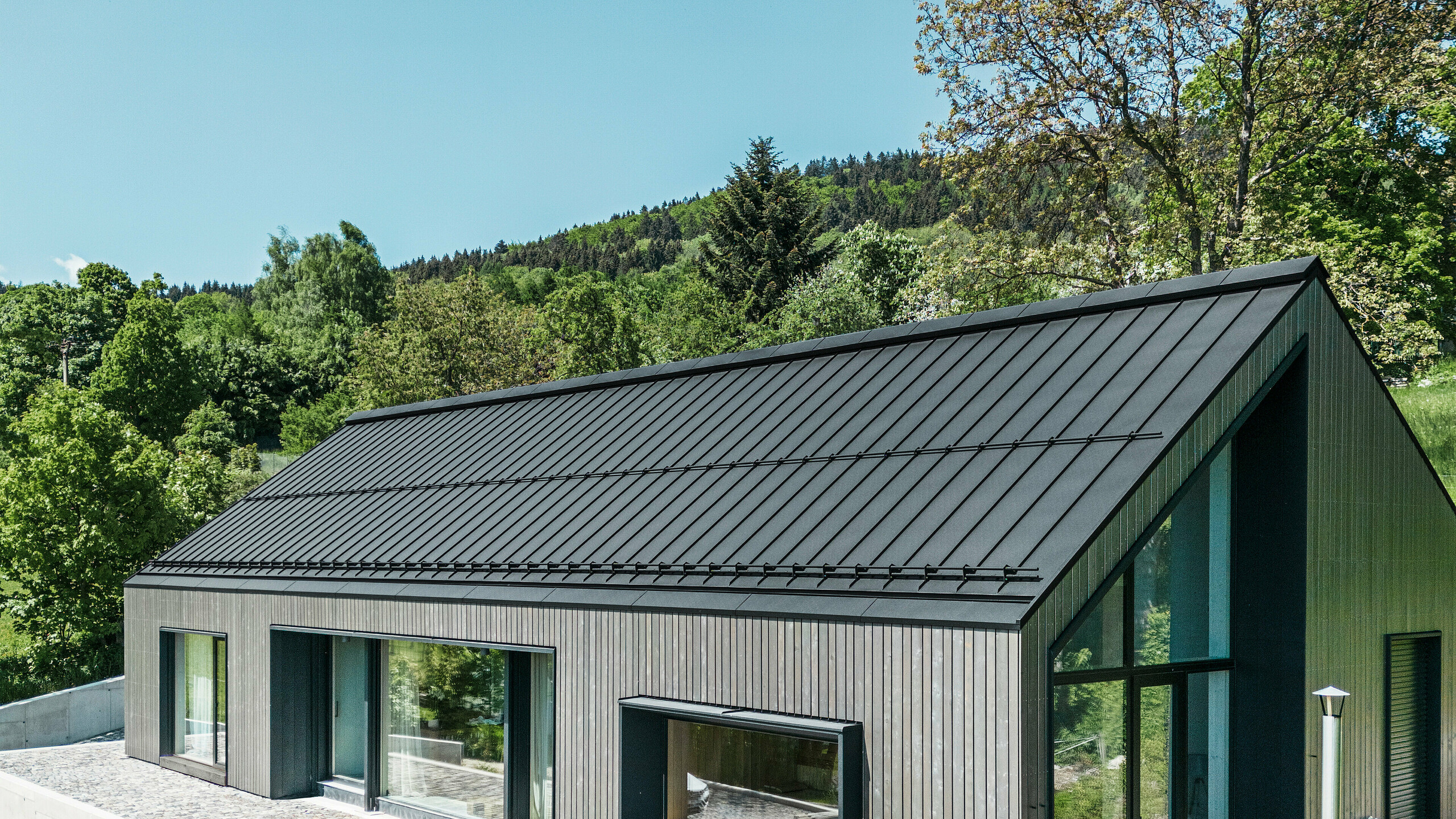 A modern wooden house in Kuncice (CZ) was equipped with large windows. The dark wooden house has a PREFALZ roof system in the colour P.10 black. In the rear outdoor area, there is a hot tub with wooden panelling.