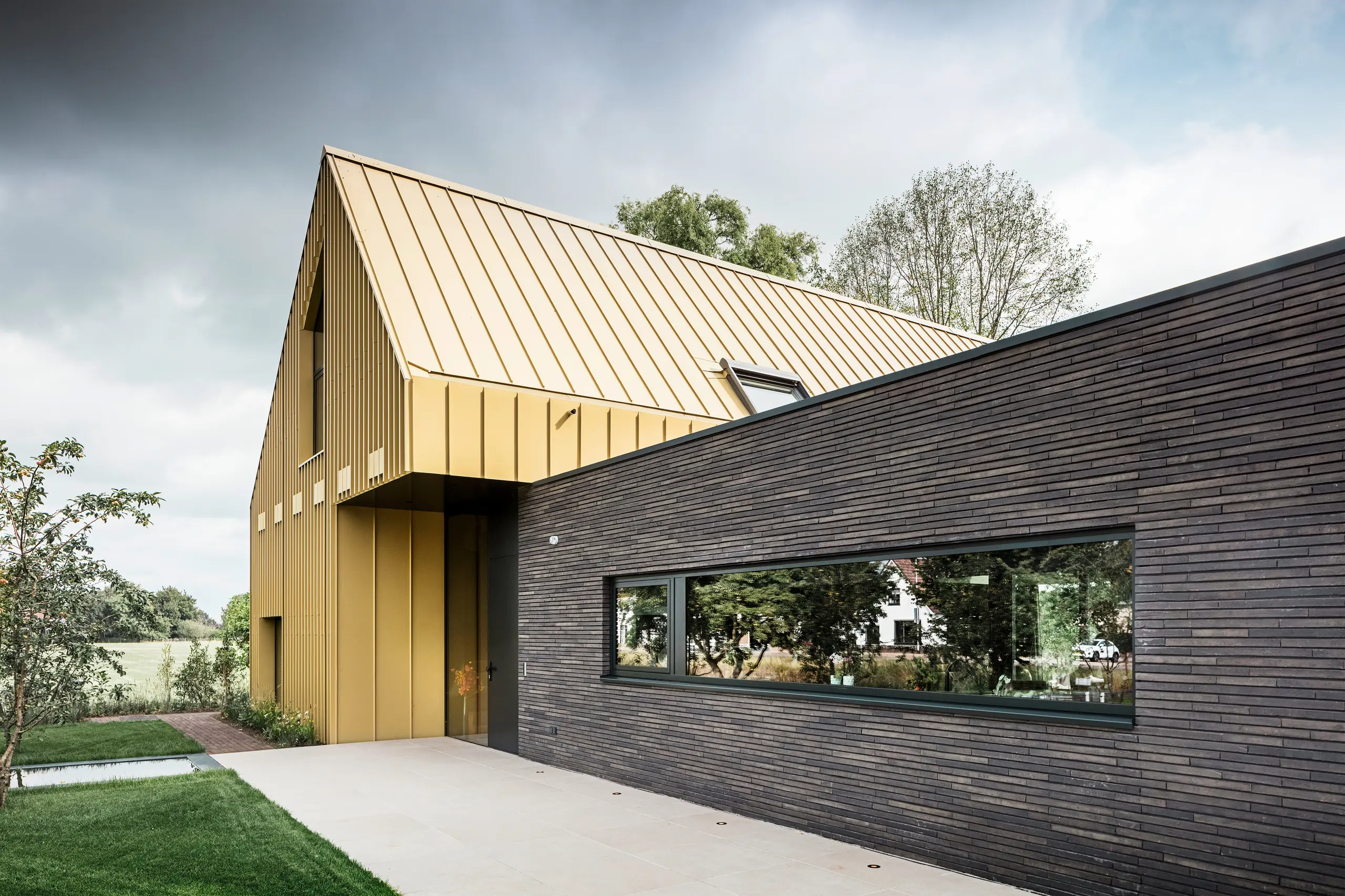 A modern detached house with a low-rise extension; the main roof and parts of the façade are clad in PREFA FALZONAL savannah beige aluminium; a large panoramic window in the extension, a surrounding garden with a lawn, trees in the background, and a cloudy sky.