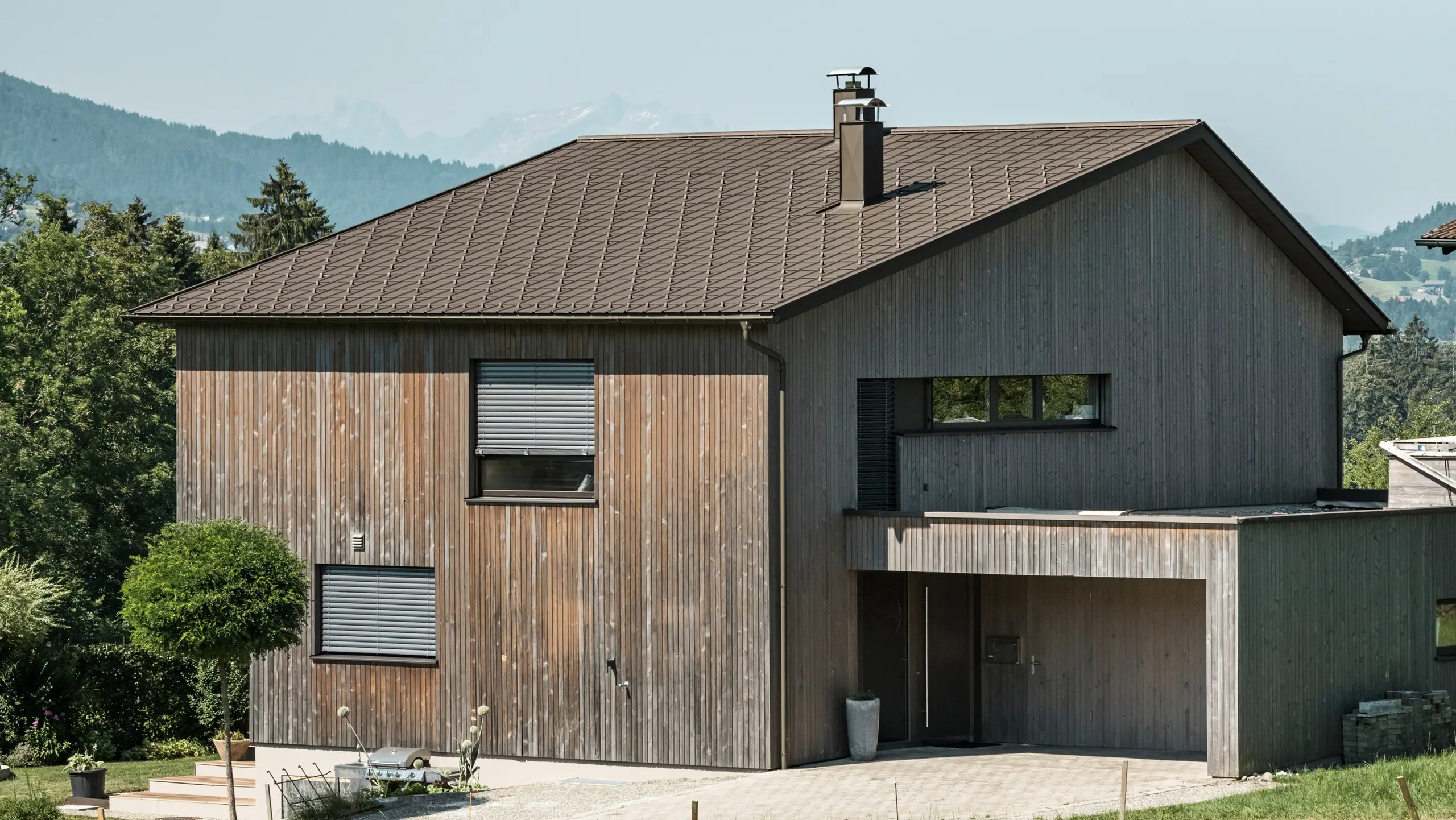 Side view of a contemporary detached house in Krumbach with a combination of a natural wooden façade and a long-lasting PREFA aluminium roof in P.10 brown. The harmonious colour scheme shows the elegant integration of modern design and traditional materials, with the PREFA roof tiles providing an aesthetic and functional roof design.