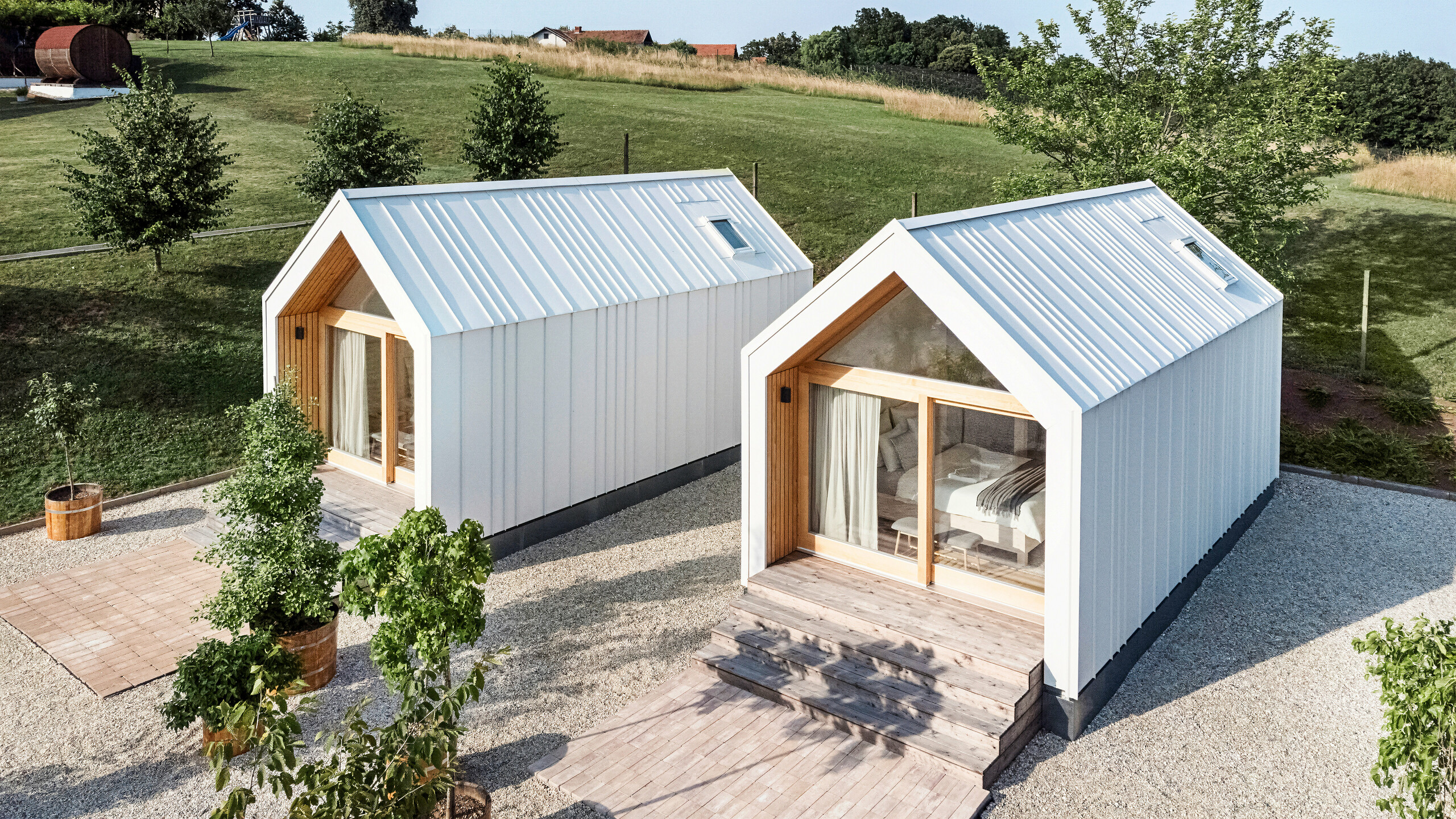 View of the front of the two tiny houses in the 'Pri Momi' holiday complex in Kančevci, Slovenia. The micro houses were built using a timber frame construction and finished with a robust PREFALZ roof and façade cladding in P.10 Pure White. The modern, minimalist buildings combine a durable yet aesthetic aluminium shell with large glass fronts and natural wood, embedded in a green landscape. The generous wooden steps lead up to the entrances, creating a cosy and inviting atmosphere.