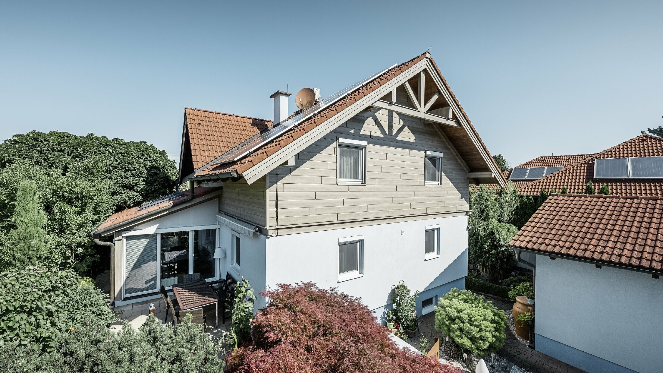 Detached house with a gable roof and red roofing, combined with a modern façade design. The upper gable is clad with beige-grey aluminium façade panels from PREFA, while the ground floor has a white plaster façade. Solar panels are installed on the roof. The house is set in a green garden with a terrace, garden furniture and lush planting, surrounded by other residential buildings with similar roof architecture.