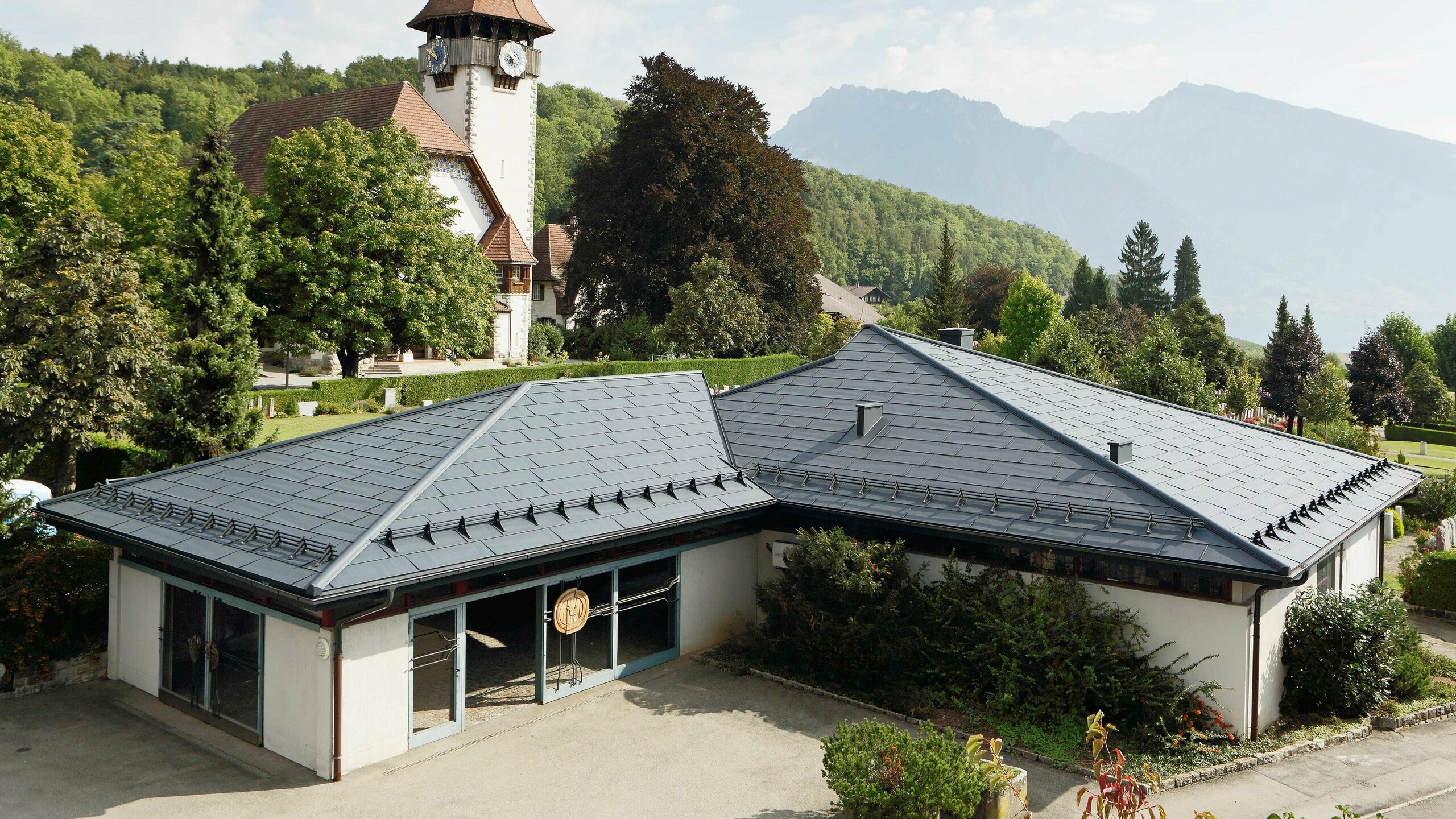 Funeral home with PREFA FX.12 roof panels in anthracite, with a church in the background and the cemetery next to it.