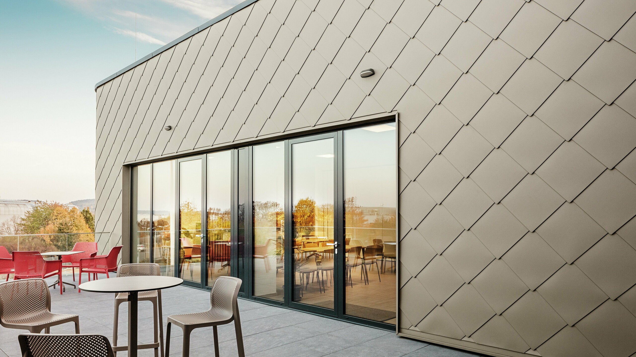 [Translate to English:] Die Fassade des neuen PREFA Standortes in Törökbálint besticht durch die eleganten PREFA Wandrauten 44 × 44 in Bronze. Die präzise verlegte Aluminiumfassade sorgt für ein modernes, dynamisches Erscheinungsbild und bietet gleichzeitig maximalen Schutz vor Witterungseinflüssen. Die großzügige Glasfront verbindet Innen- und Außenbereich und schafft auf der großzügigen Terrasse eine helle und einladende Atmosphäre.