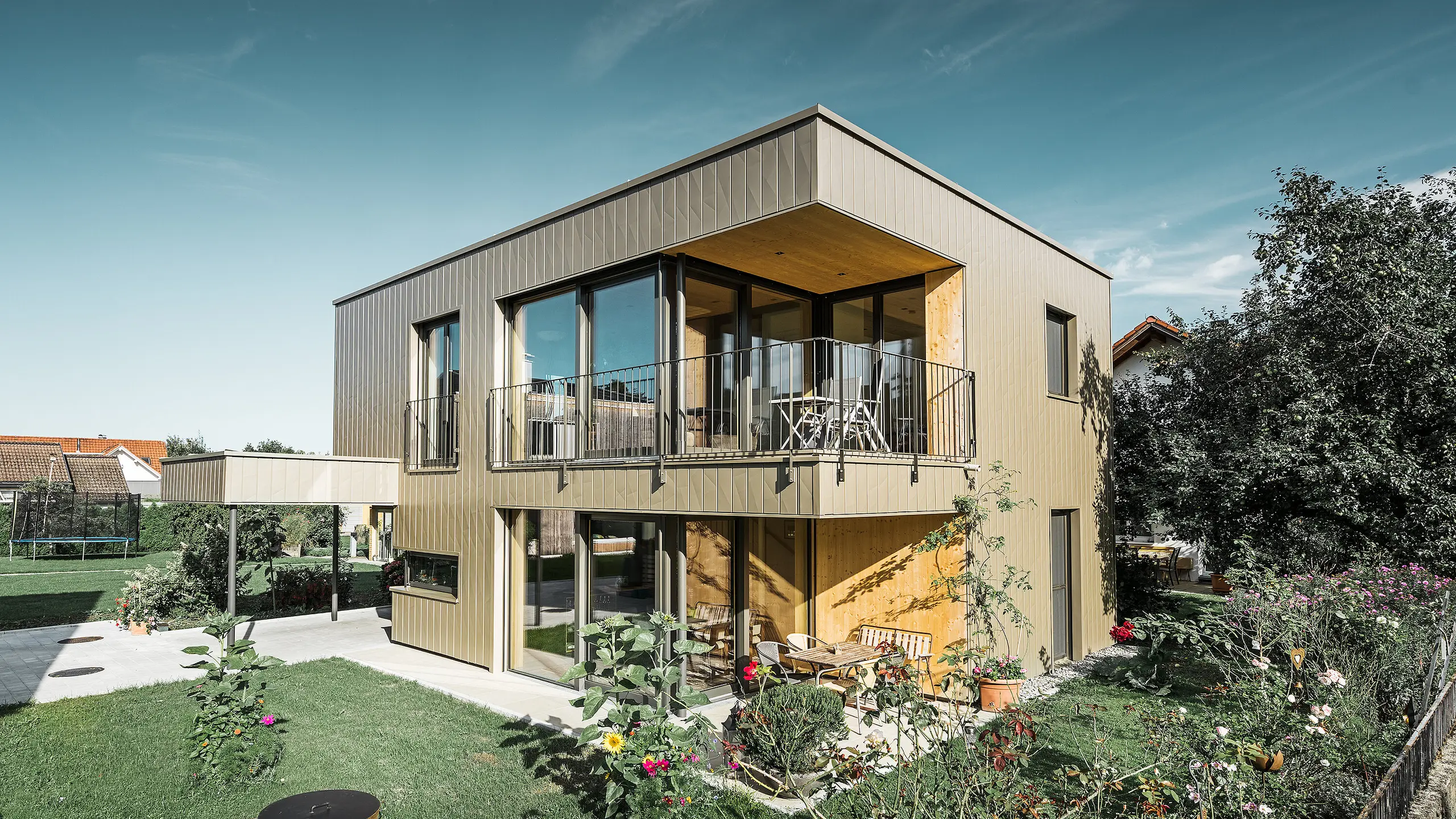 Moderner Neubau mit Flachdach und großen Fensterflächen; Der Großteil der Fassade ist mit vertikalen Alu-Fassaden-Paneelen von PREFA, Siding.X, in Bronze verkleidet. Man erhält auch einen Blick auf den rundum liegenden Garten.
