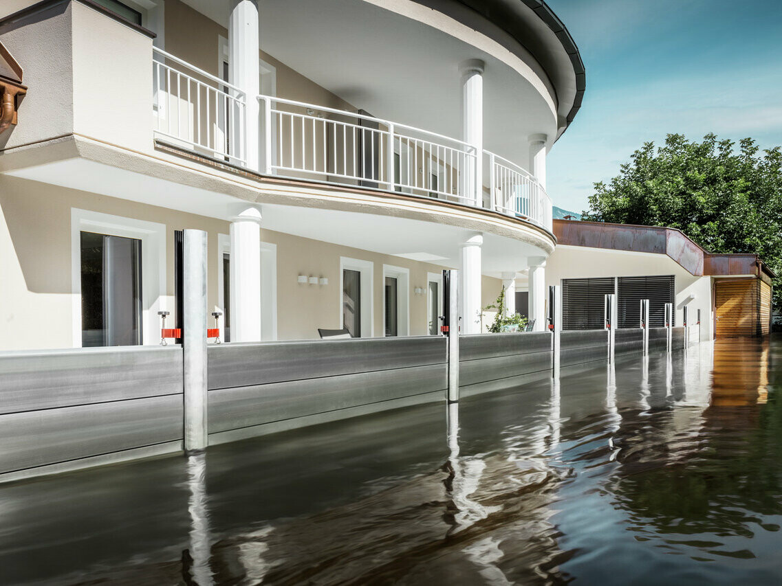 The picture shows a detached house with a balcony. The PREFA flood protection system protects the house from the already rising flood waters.