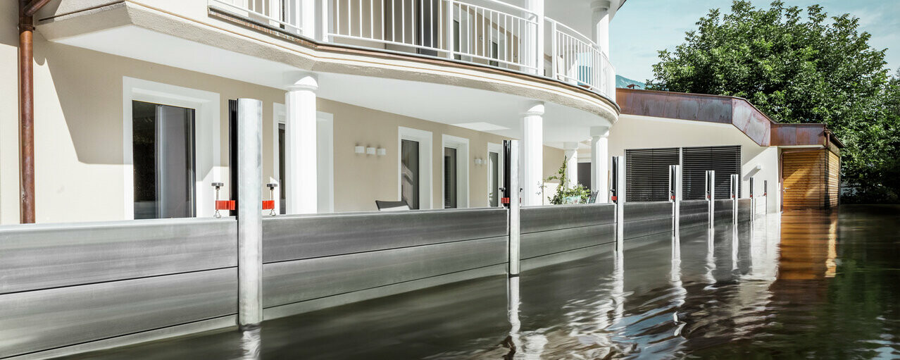 The picture shows a detached house with a balcony. The PREFA flood protection system protects the house from the already rising flood waters.